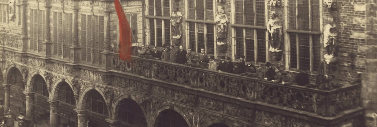 Das Bild zeigt eine historische, großangelegte Versammlung auf einem öffentlichen Platz, der von einer großen Menschenmenge bedeckt ist, die nach oben zu einem balkonartigen Vorsprung eines gotischen Gebäudes blickt. Auf dem Balkon, der mit Bögen und reicher Verzierung gestaltet ist, befinden sich mehrere Personen, die offensichtlich eine wichtige Rede oder Veranstaltung beobachten oder leiten. Markant ist eine große rote Fahne, die auf der linken Seite des Balkons weht und einen starken visuellen Akzent setzt.  