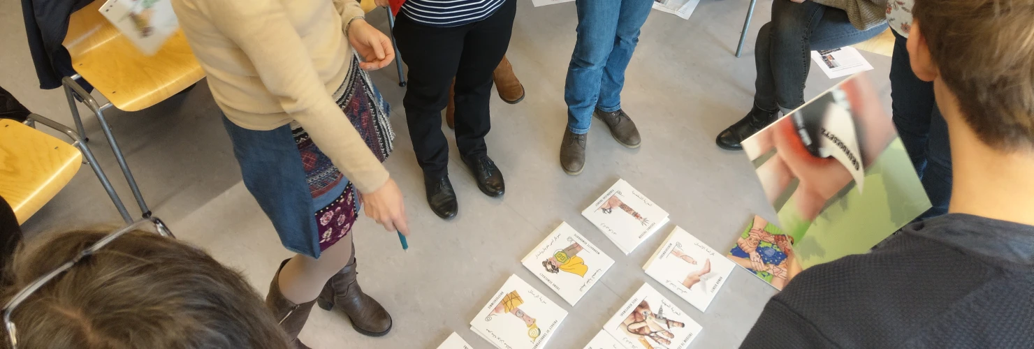 Das Bild zeigt eine Gruppe von sieben Personen, die in einem Klassenzimmer in einem Kreis stehen und sich auf Spielkarten konzentrieren, die vor ihnen ausgebreitet sind.   