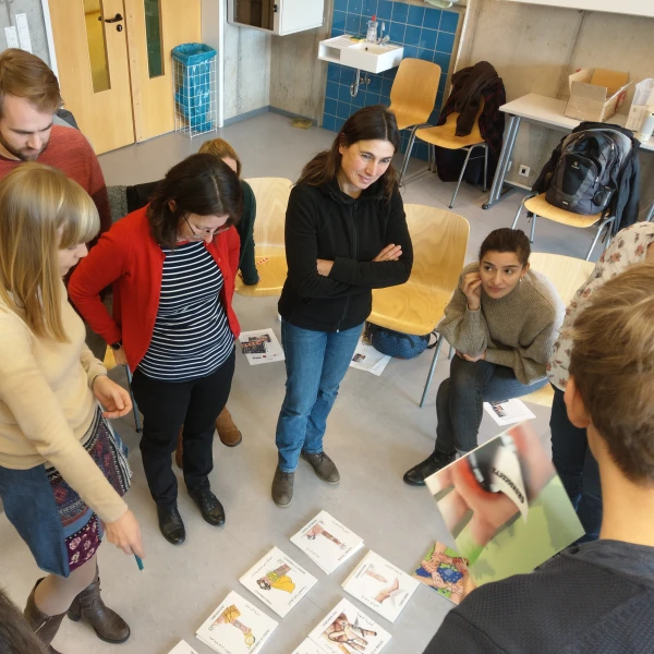 Das Bild zeigt eine Gruppe von sieben Personen, die in einem Klassenzimmer in einem Kreis stehen und sich auf Spielkarten konzentrieren, die vor ihnen ausgebreitet sind.   