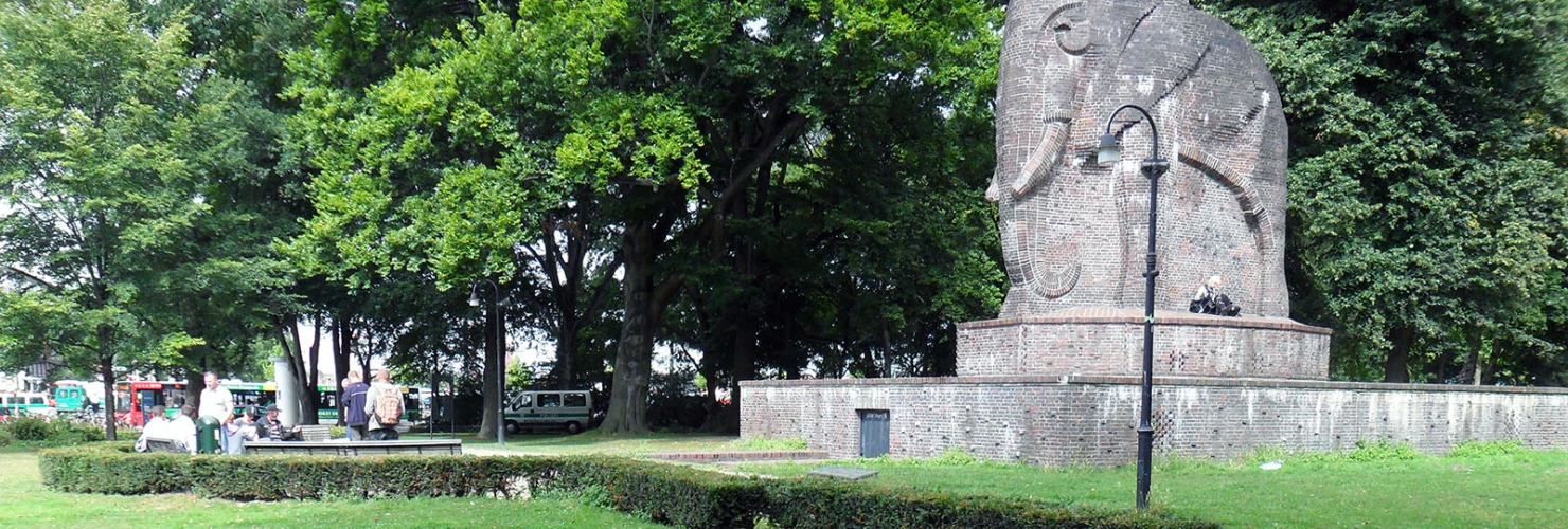 Das Bild zeigt einen Park mit einem großen Elefanten-Steindenkmal in einer Naturumgebung. Im Vordergrund befindet sich ein kreisförmiges Beet, welches mit vielen kleinen und großen Steinen gefüllt ist. Der Hintergrund wird von grünen Bäumen und einem gepflasterten Weg, der durch den Park führt, geprägt.