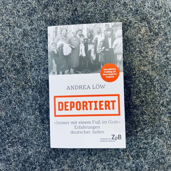 Auf dem Bild ist das Cover eines Buches mit dem Titel "Deportiert" von Andrea Löw zu sehen. Das Cover zeigt oben ein schwarz-weiß Foto von einer Gruppe lächelnder Menschen und darunter den Buchtitel in großer, orangener Schrift auf einem grauen Hintergrund, umrahm von einem orangenen Kasten. Der Untertitel „Immer mit einem Fuß im Grab: Erfahrungen deutscher Juden“ ist in grauer Schrift direkt unter dem Haupttitel angebracht.  